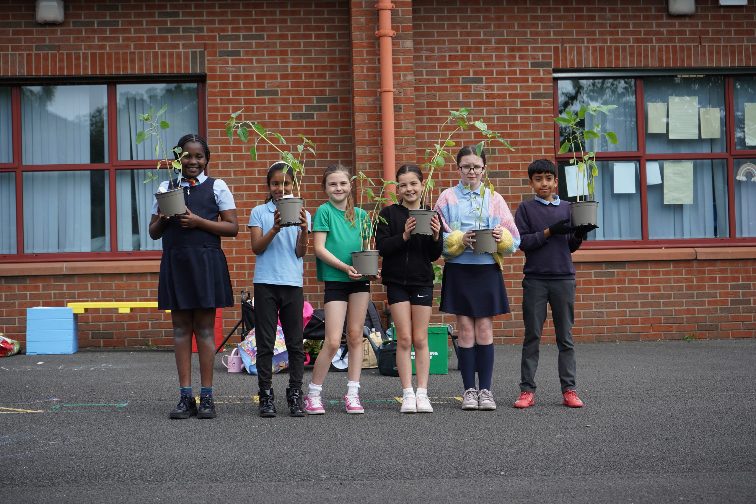 six children, standing in front of a red brick building, holding flower pots